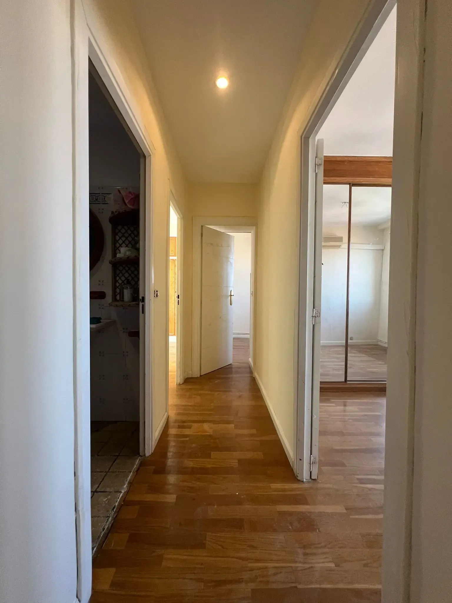 Pasillo estrecho con suelos de madera, paredes blancas, luces de techo y puertas abiertas a las habitaciones a ambos lados.