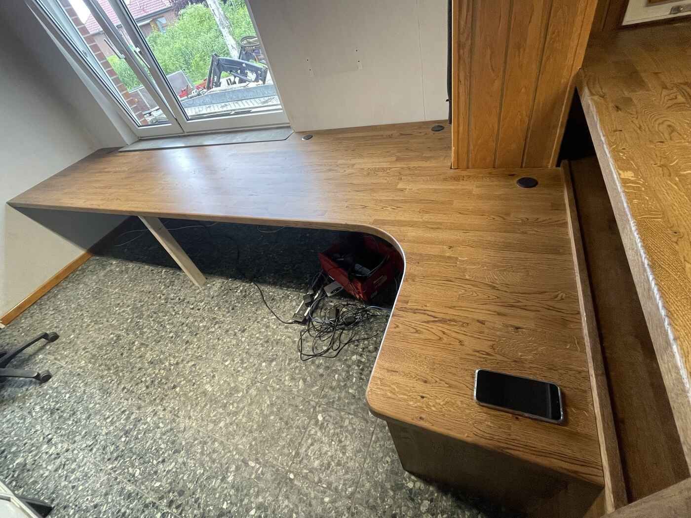 Ein Holzschreibtisch mit einem Mobiltelefon darauf in einem Raum neben einem Fenster.