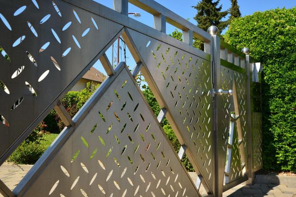 Zäune, Tore & Sichtschutz Modernes graues Metalltor mit geometrischen Ausschnitten neben einer grünen Hecke und einer gepflasterten Auffahrt