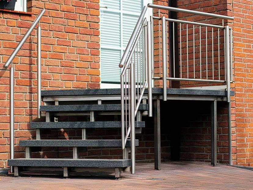 Graue Metalltreppe und Plattform neben dem Eingang eines Backsteingebäudes mit Handläufen.