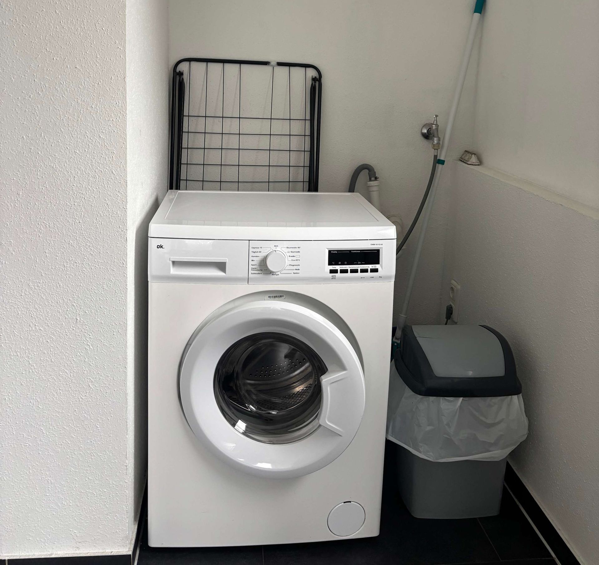 Eine weiße Waschmaschine steht in einer Waschküche neben einem Mülleimer