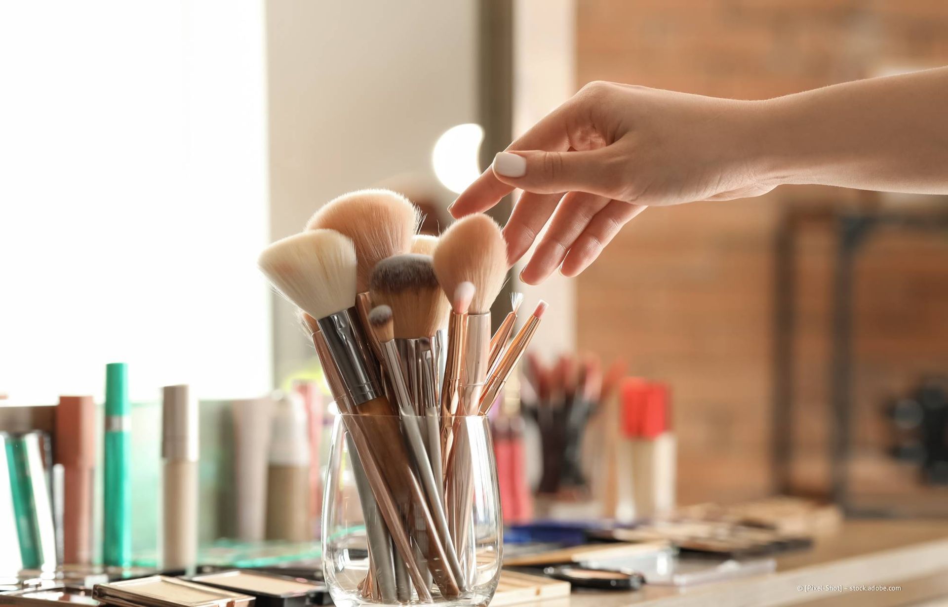 Hand greift nach einem Make-up-Pinsel in einem Glas. Weitere Pinsel, Kosmetika auf einem Regal. Helle Umgebung.