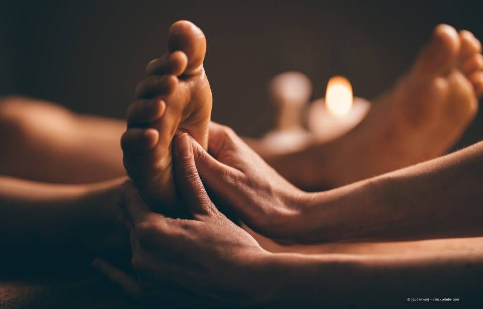 Hände massieren die Fußsohle vor einem unscharfen Hintergrund mit Kerzen.