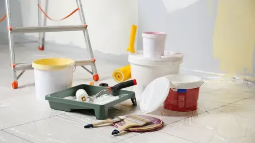 En el suelo de una habitación hay cubos de pintura, rodillos, pinceles y una escalera.