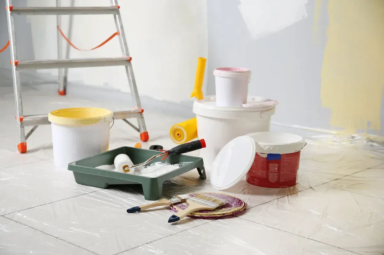 Una habitación siendo pintada con baldes de pintura, rodillos, pinceles y una escalera.