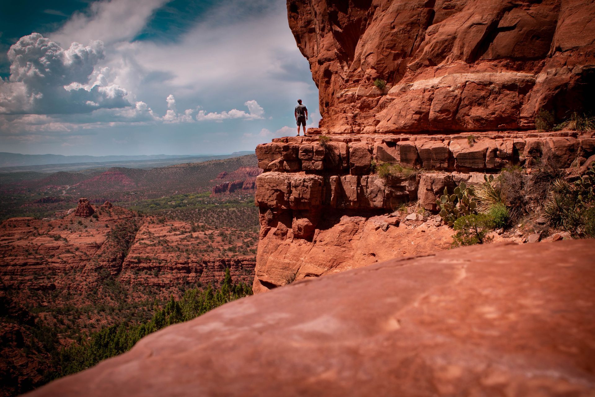 Person standing on a red rock ledge overlooking a canyon under a cloudy sky
