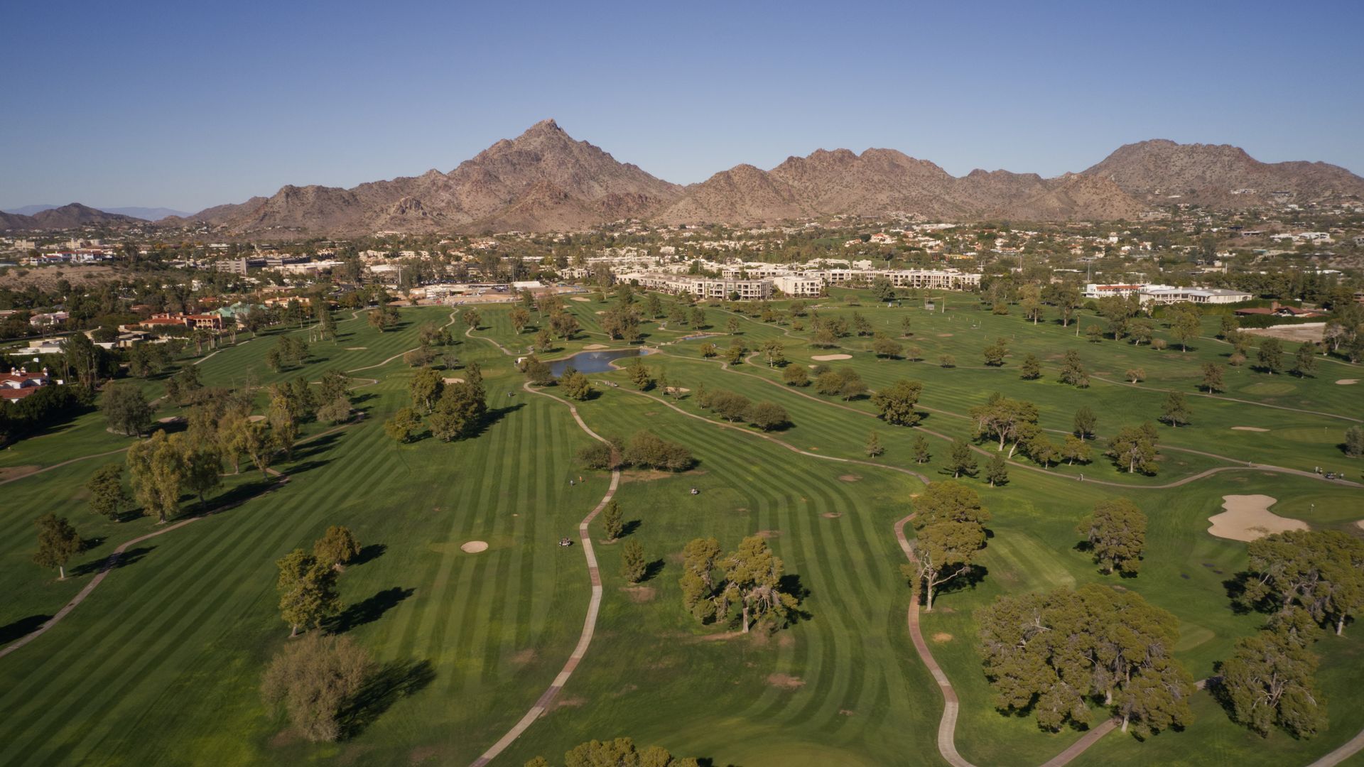 Wide aerial view of a green golf course with scattered trees and desert mountains in the background