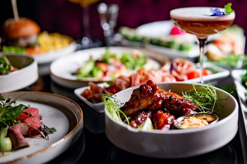Plated gourmet dishes and a cocktail on a restaurant table, with colorful appetizers in the background