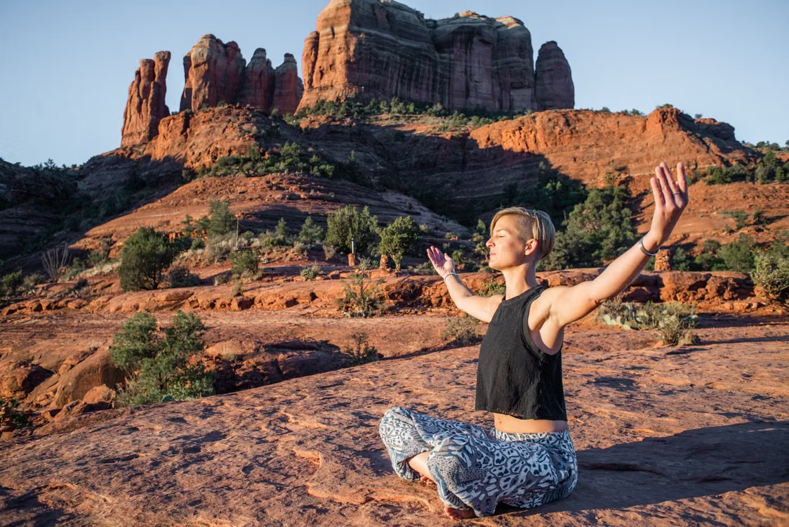 Woman meditating on a rocky desert cliff with arms raised, red sandstone mesas in the background
