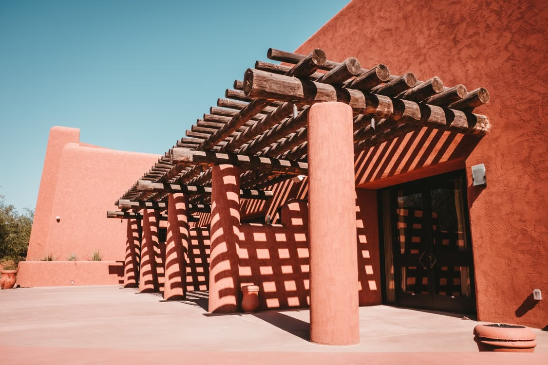 Adobe building with pink stucco walls and wooden pergola shadows in a sunlit courtyard.