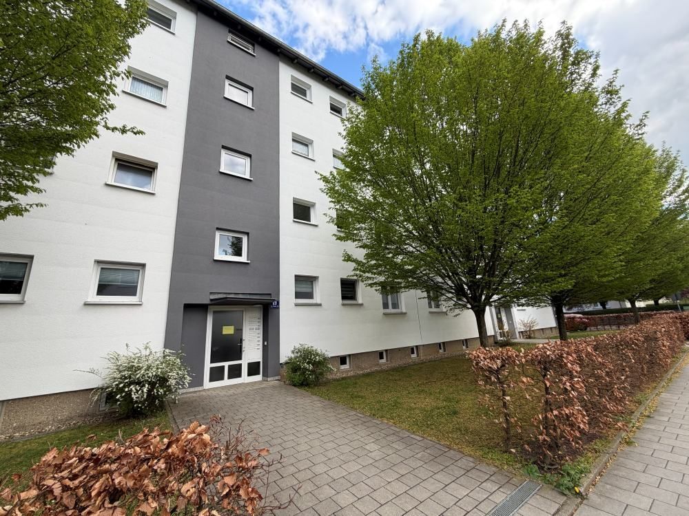 Eingang zum Wohnhaus mit grau-weißer Fassade, gepflastertem Gehweg, Bäumen und herabgefallenen Blättern
