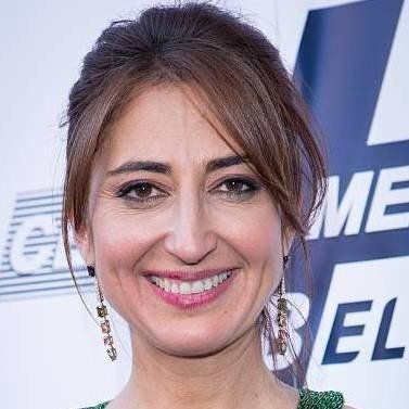 Mujer con cabello castaño sonriendo a la cámara, con aretes y top verde, frente a un fondo borroso.