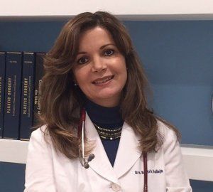 Mujer con bata blanca, estetoscopio, sonriente, cabello castaño, cuello alto azul, entorno de oficina.