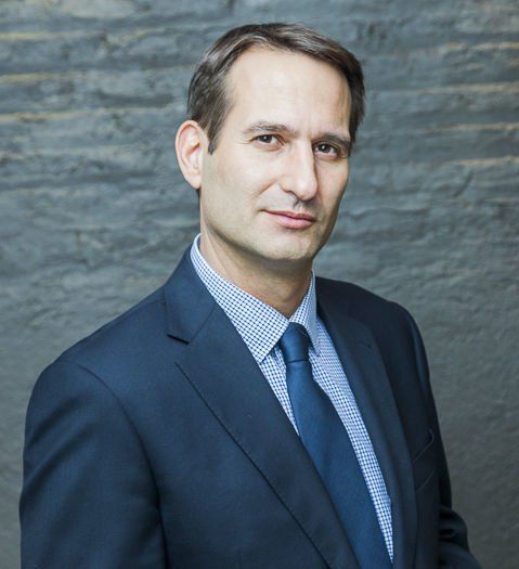 Hombre con traje azul y corbata, mirando a la cámara, de pie frente a una pared de ladrillos grises.