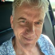 Hombre de cabello gris, sonriendo en un automóvil, vistiendo una camisa estampada azul.