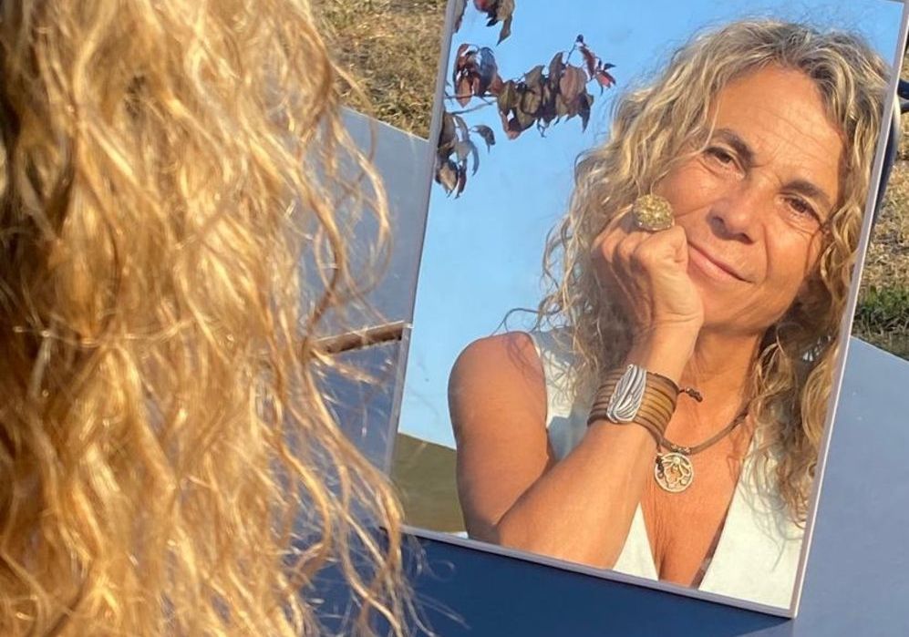 Una mujer con cabello rubio rizado se mira en el espejo al aire libre, sonriendo, luciendo joyas y una blusa blanca.