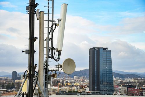 Antenas de telefonía móvil y una antena parabólica en primer plano, con el perfil de la ciudad y las montañas bajo un cielo azul nublado.