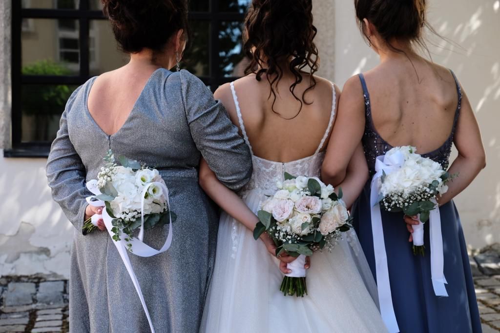 Braut und Brautjungfern mit Blumensträußen, mit dem Rücken zur Kamera. Brautjungfern in grauen und blauen Kleidern stehen neben einem weißen Gebäude.