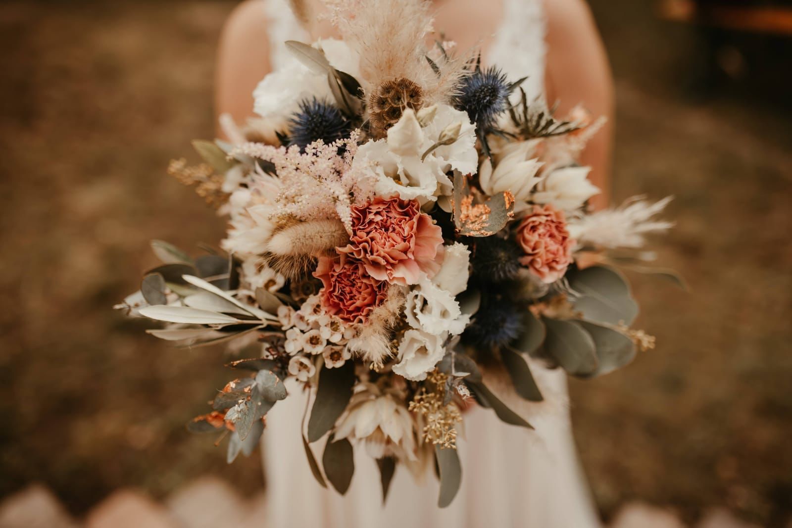 Braut hält einen Strauß pfirsichfarbener, weißer und blauer Blumen mit Grün.