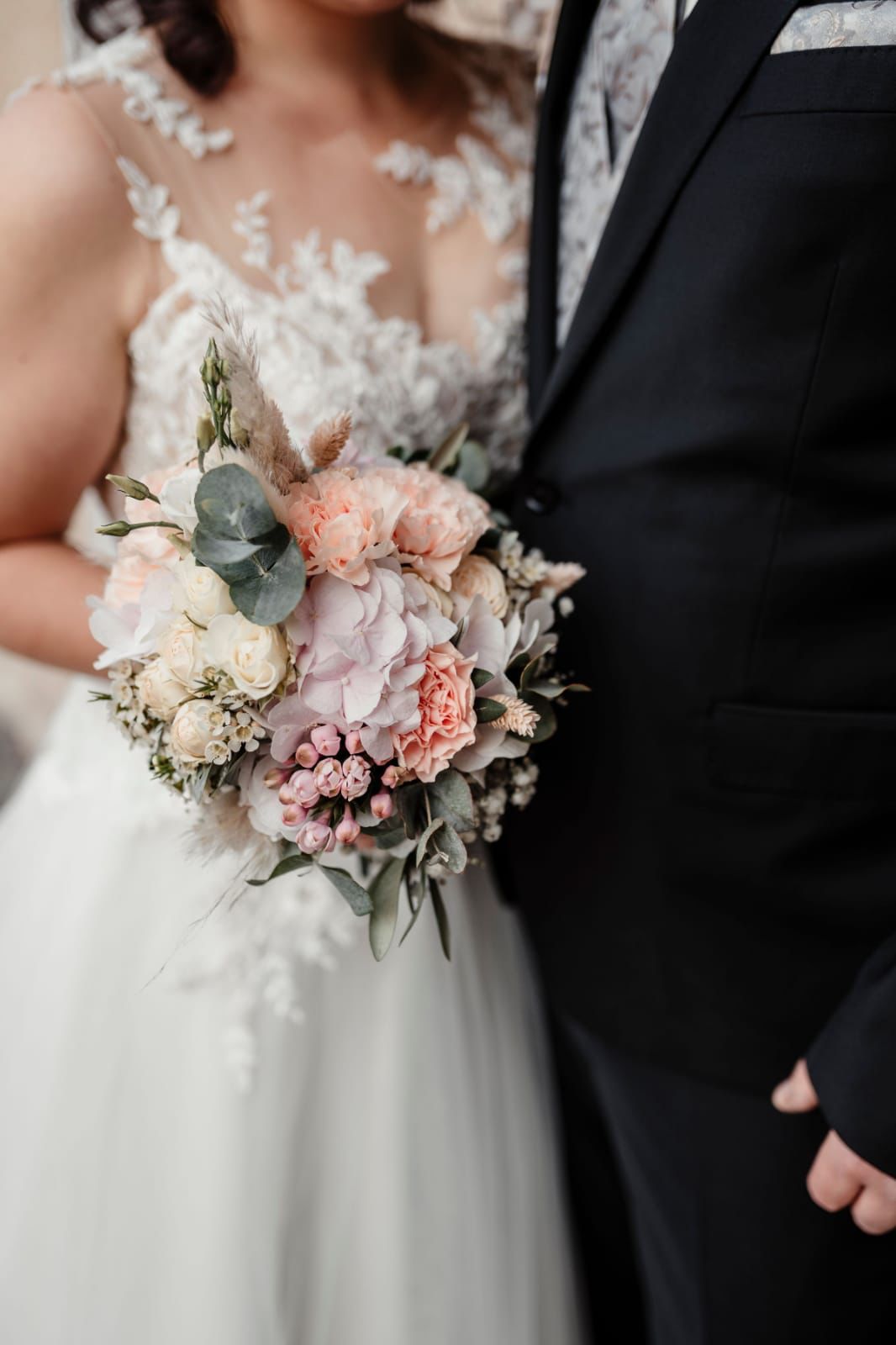 Braut und Bräutigam halten einen Blumenstrauß in der Hand, die Braut trägt ein weißes Kleid, der Bräutigam einen schwarzen Anzug.