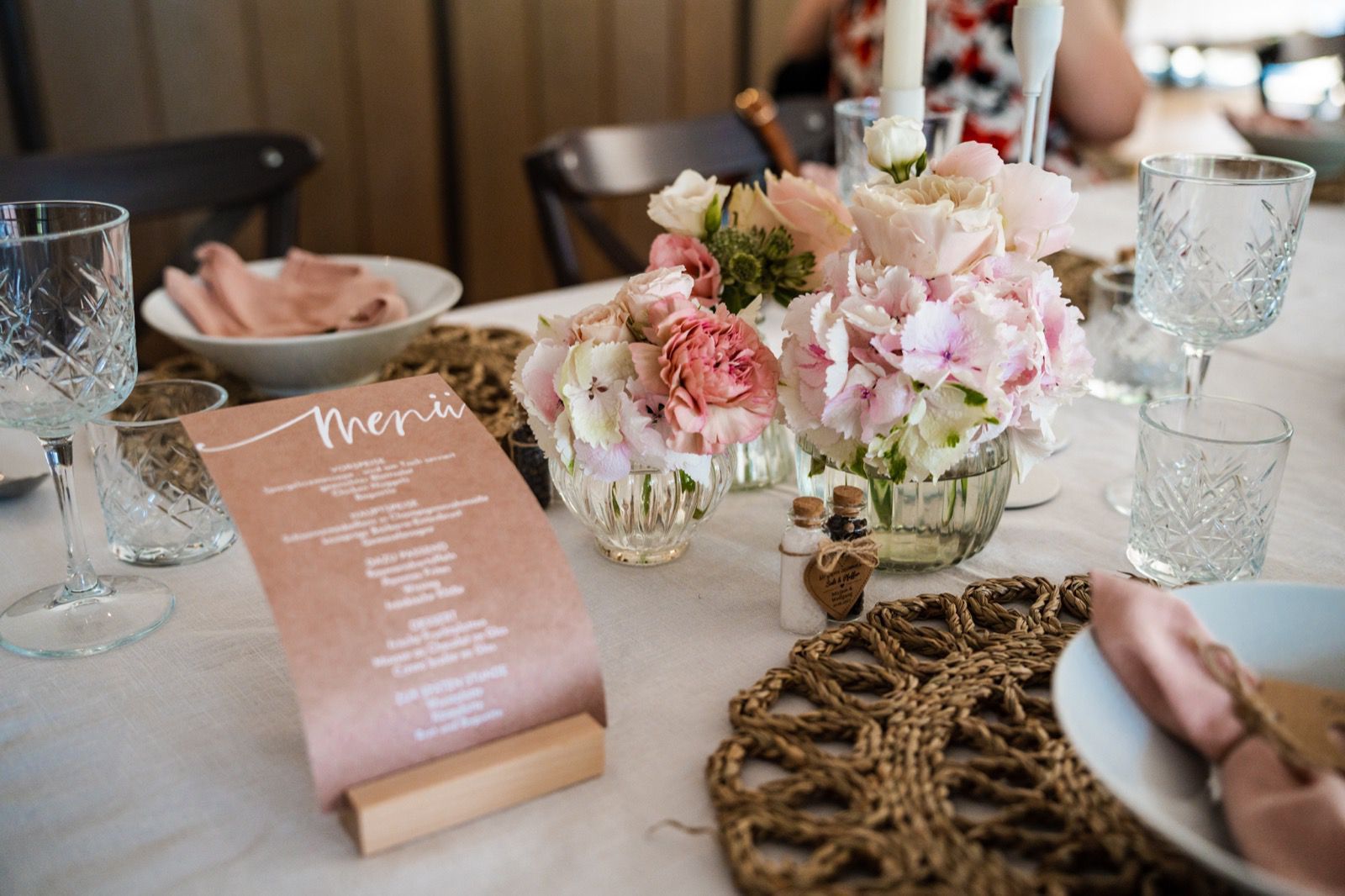 Tischdekoration: Menükarte, Blumenschmuck, Gedecke mit rosa Akzenten.