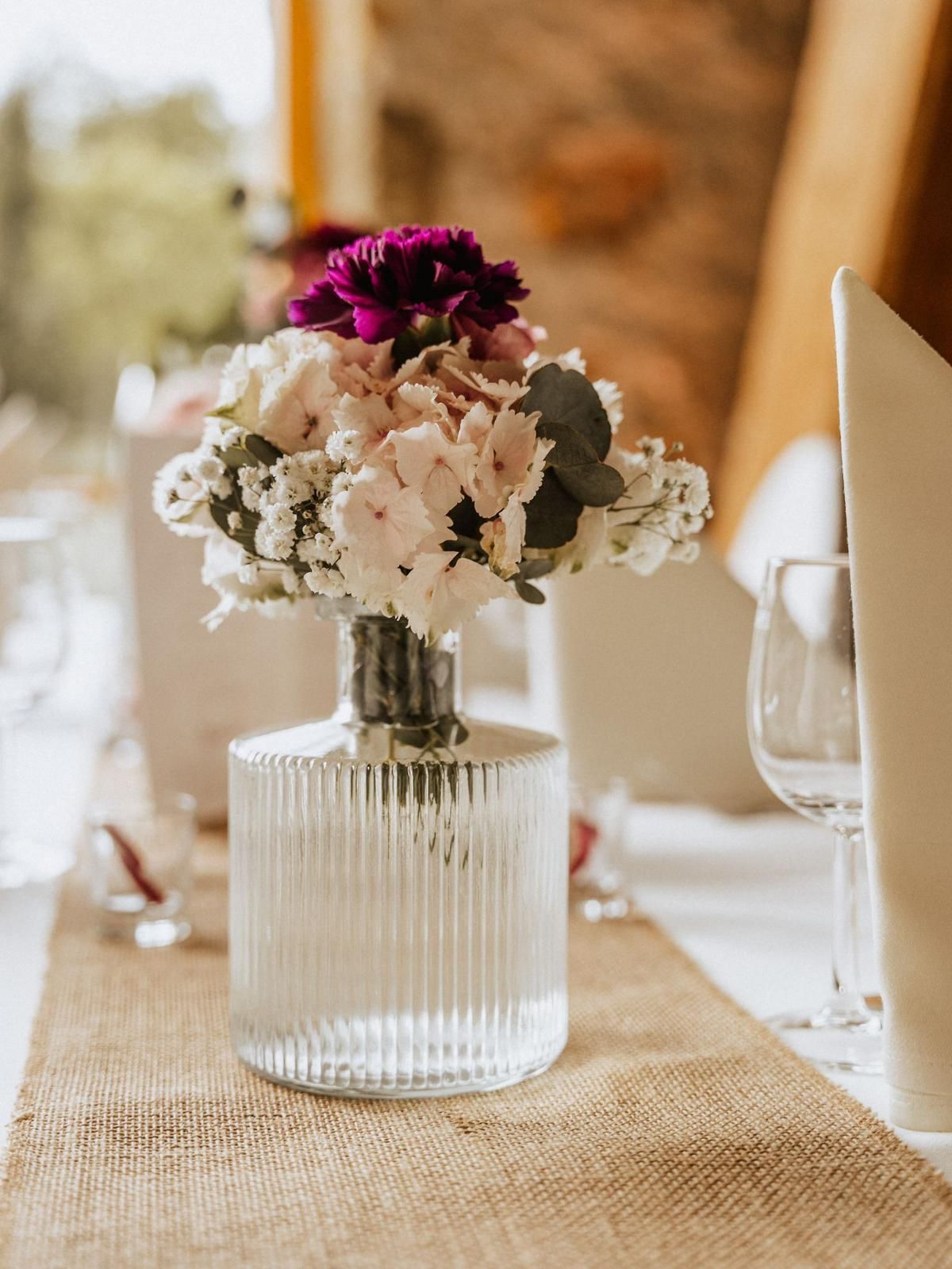 Blumenarrangement in einer durchsichtigen, gerippten Vase auf einem Tischläufer aus Sackleinen.