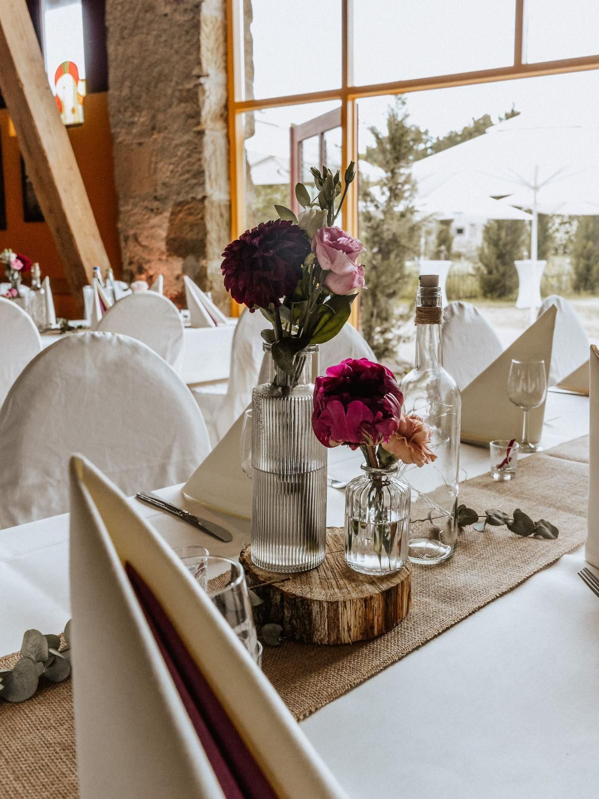 Hochzeitstisch mit Blumen, Weinflasche und Servietten auf einem Juteläufer. Heller Raum mit einem Fenster.
