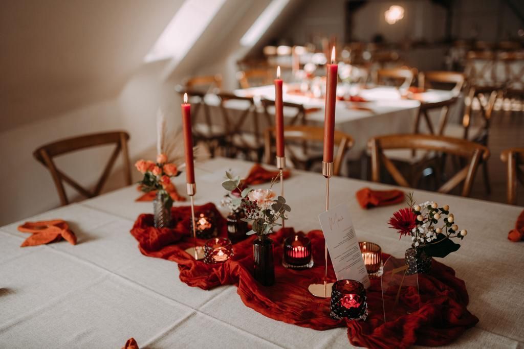 Hochzeitsempfangstisch mit roten Kerzen und Läufer, weißer Tischdecke, Holzstühlen.