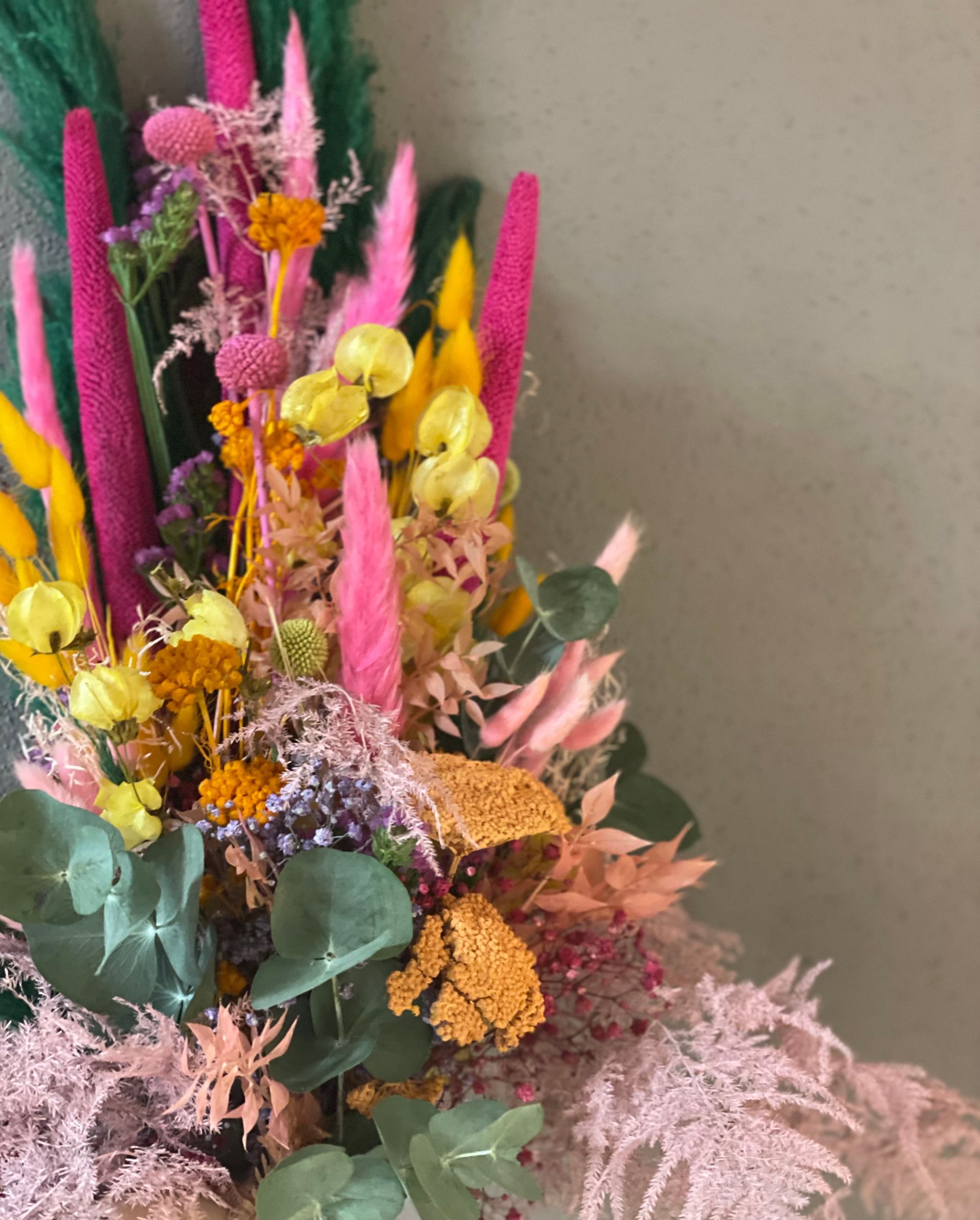 Farbenfrohes Trockenblumenarrangement vor grünem Hintergrund mit rosa, gelben und orangefarbenen Farbtönen.
