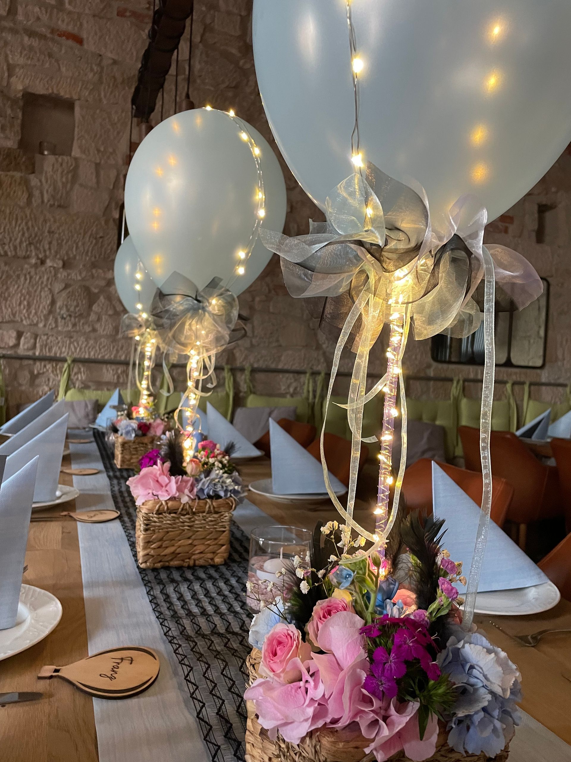 Tischdekoration: Beleuchtete Ballons auf Blumenarrangements, aufgestellt auf einem langen Tisch in einem gut beleuchteten Raum.