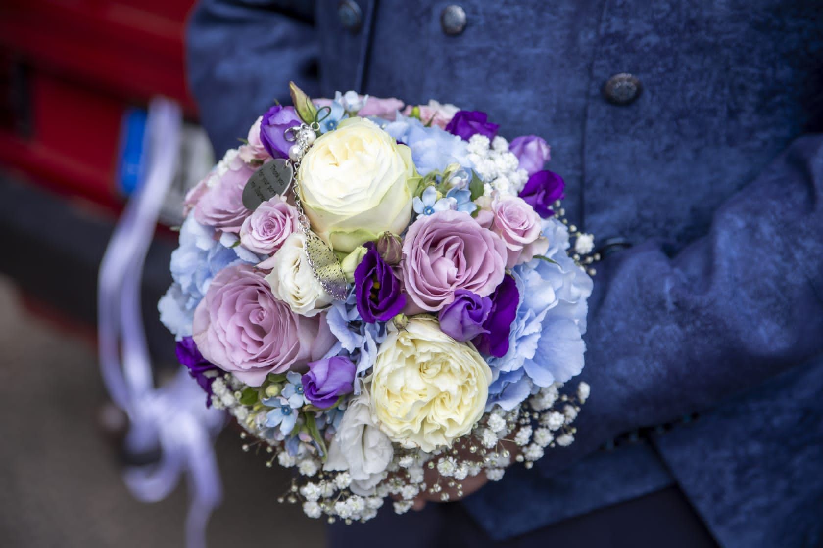 Nahaufnahme eines Straußes aus pastellfarbenen Rosen, blauen Hortensien und violetten Blumen, der von einer Person in einer blauen Jacke gehalten wird.