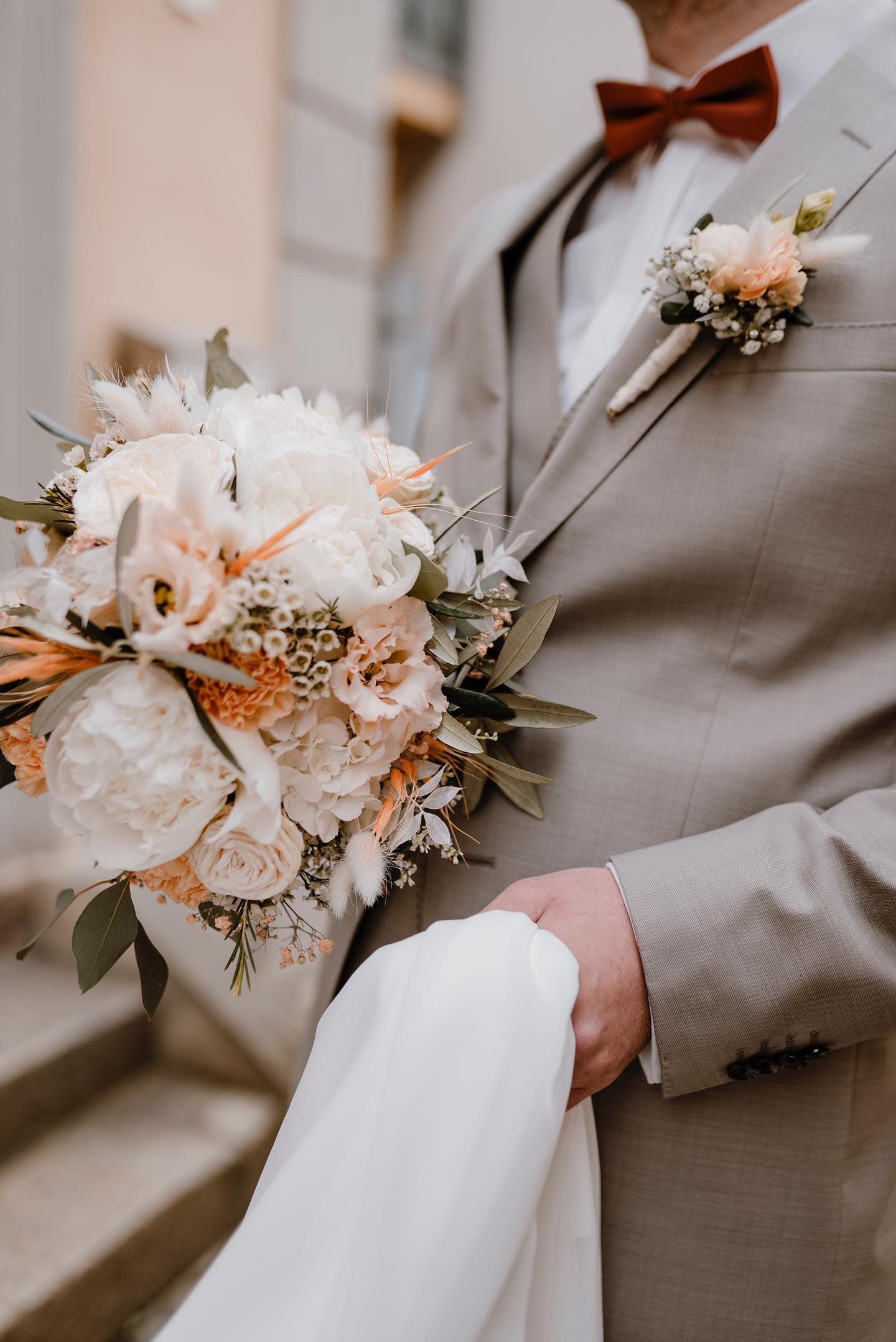 Bräutigam hält Blumenstrauß und Schleier in der Hand, trägt einen grauen Anzug mit roter Fliege, draußen.