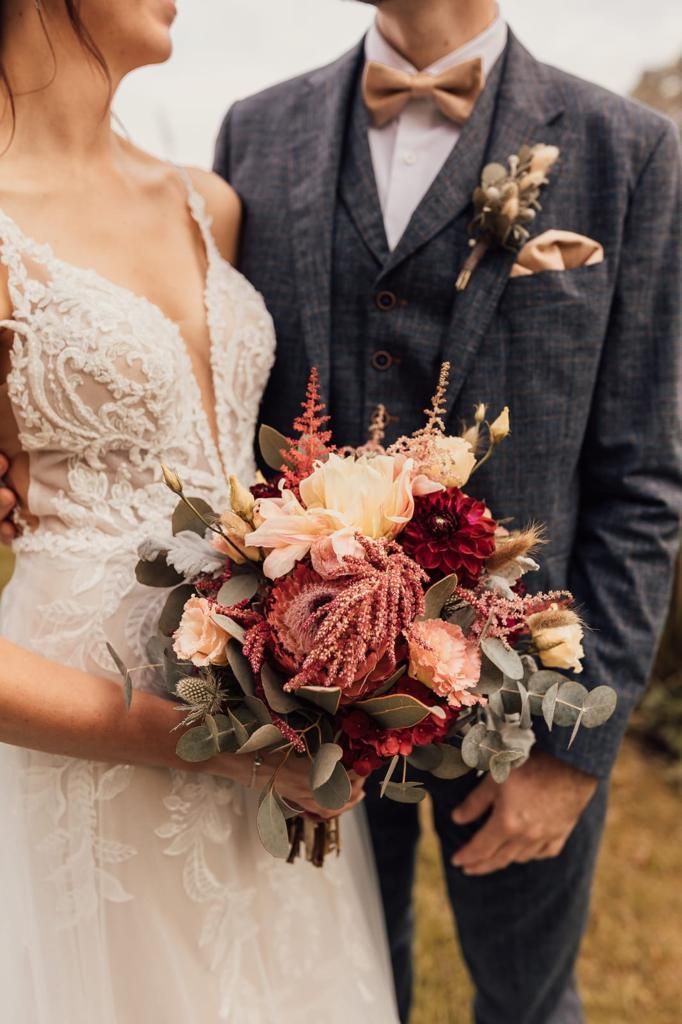 Braut und Bräutigam umarmen sich und halten einen Blumenstrauß in den Händen. Die Braut trägt ein weißes Spitzenkleid und der Bräutigam einen blauen Anzug.