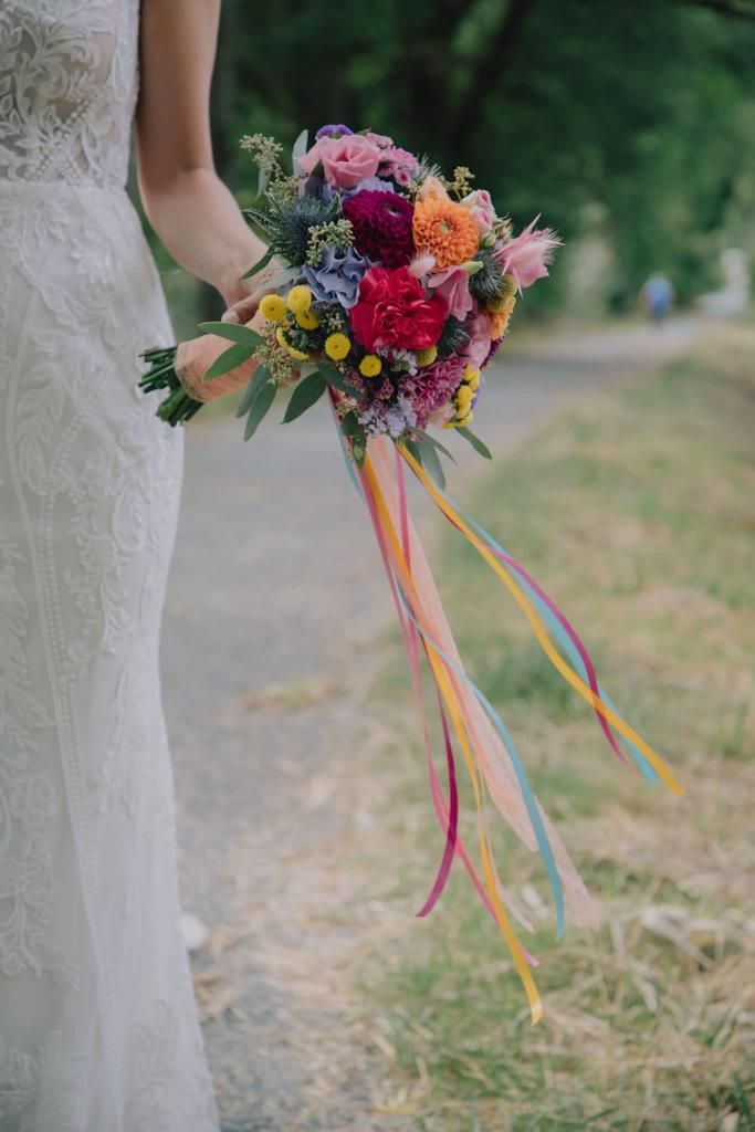 Braut im Spitzenkleid hält im Freien einen bunten Blumenstrauß mit langen Regenbogenbändern.
