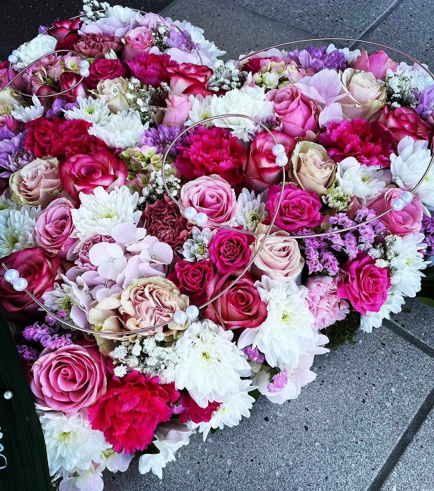 Herzförmiges Blumenarrangement mit rosa, roten und weißen Blüten auf grauem Untergrund.