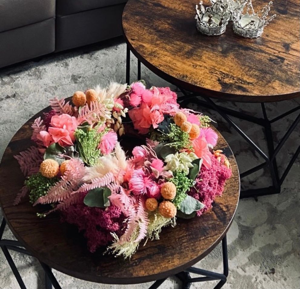 Kranz aus rosa, orangefarbenen und grünen Blumen auf einem dunklen Holztisch; zwei silberne Kronen liegen auf einem zweiten Tisch.