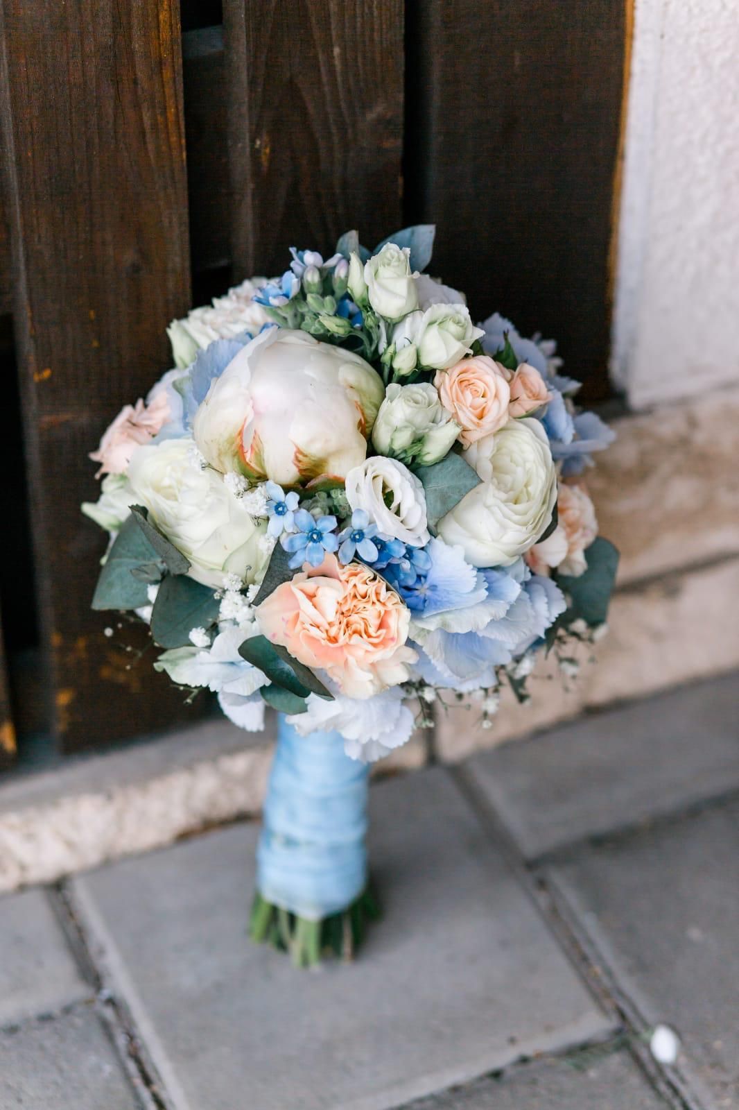 Brautstrauß aus hellblauen, pfirsichfarbenen und weißen Blumen mit einem mit einem Band umwickelten Griff auf einer Steinoberfläche.