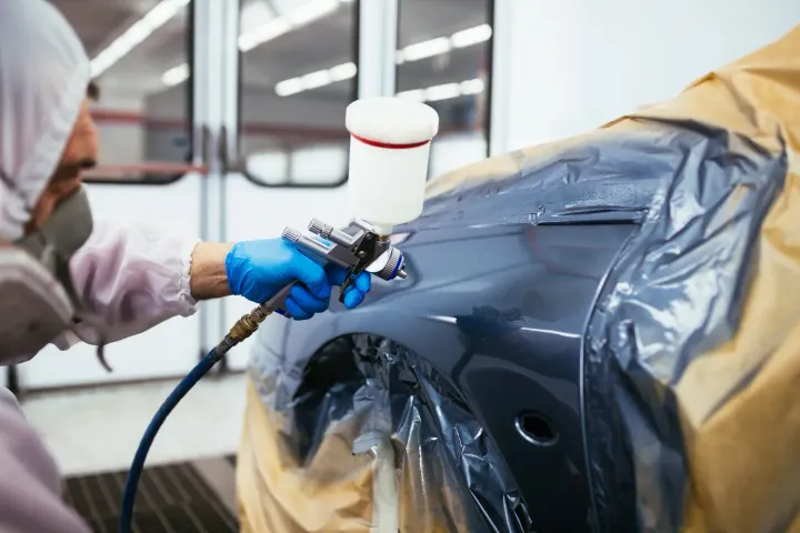Eine Person in Schutzkleidung besprüht in einer Werkstatt einen Kotflügel mit Farbe.