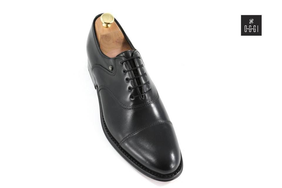 Une paire de chaussures en cuir noir avec un porte-chaussures en bois sur fond blanc.