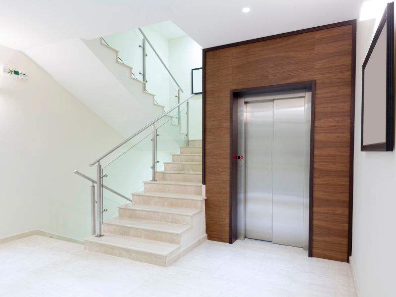 Escalera y ascensor en el vestíbulo de un edificio moderno. Paredes blancas, escaleras de mármol, barandilla de cristal y entrada de ascensor con paneles de madera.