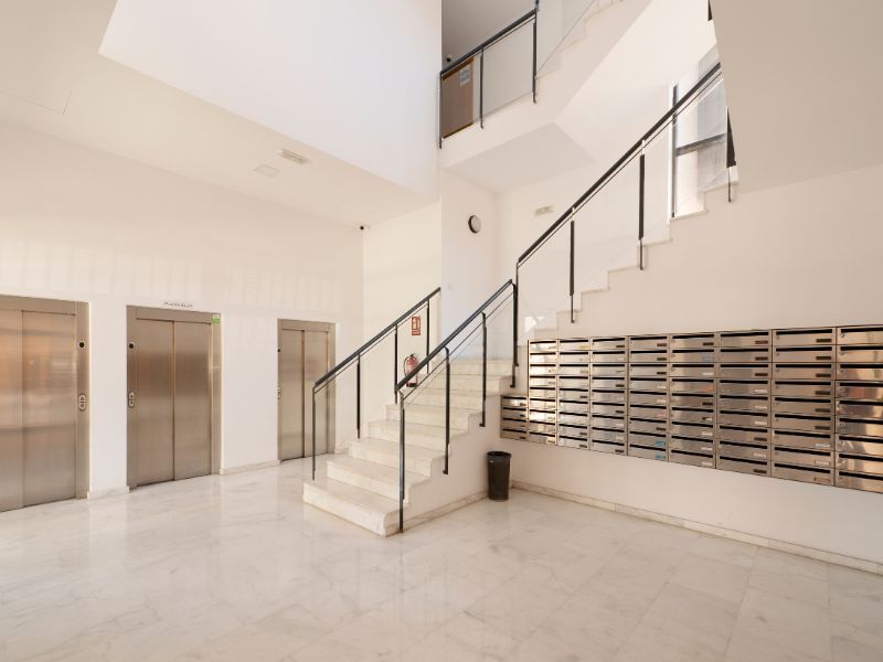 Un vestíbulo moderno y luminoso con ascensores, escaleras y buzones. Los detalles de acero inoxidable contrastan con las paredes y el suelo blancos.