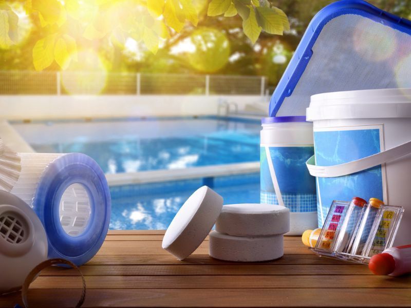 Suministros para piscina en una terraza de madera junto a una piscina. Incluye filtros, pastillas de cloro, tiras reactivas y un skimmer.