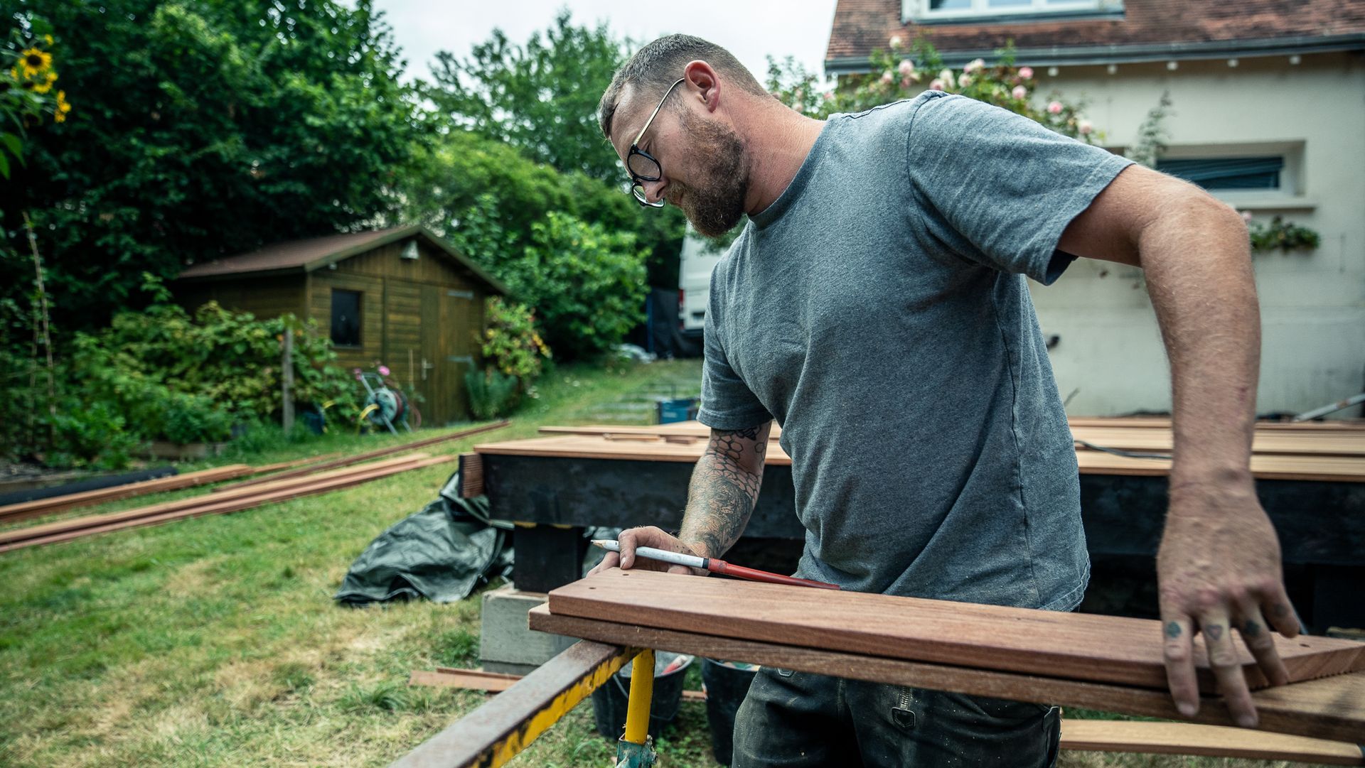 Un homme portant des lunettes travaille sur du bois à l'extérieur ; arbres verts et hangar en arrière-plan.