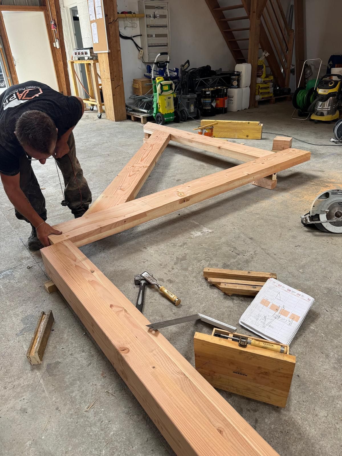 Un homme construit une structure en bois à l'intérieur. Il est concentré, assemblant des poutres. Outils et matériaux sont éparpillés au sol.