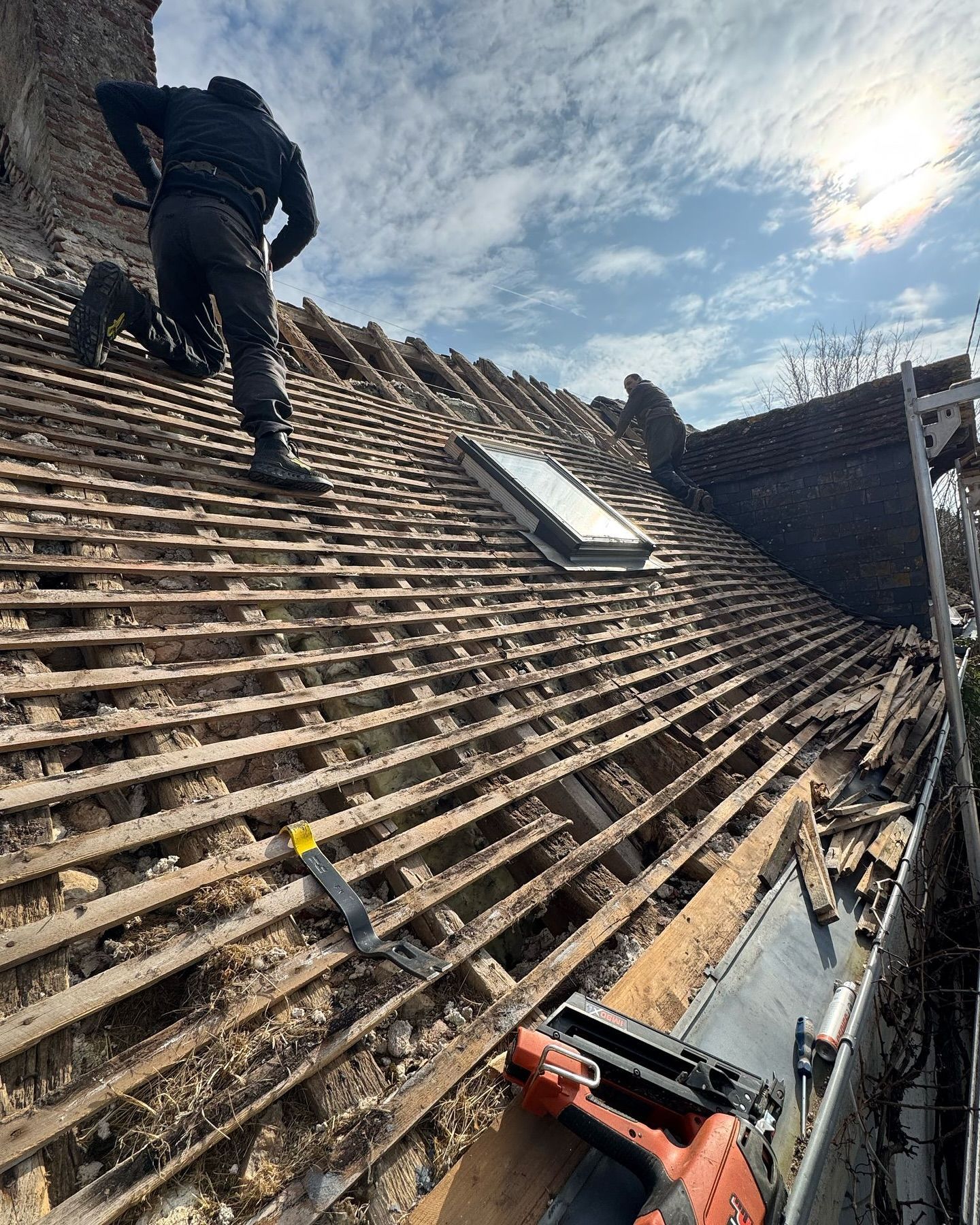 Une personne travaille sur un toit partiellement démoli. On aperçoit le ciel, le soleil et les briques du bâtiment.