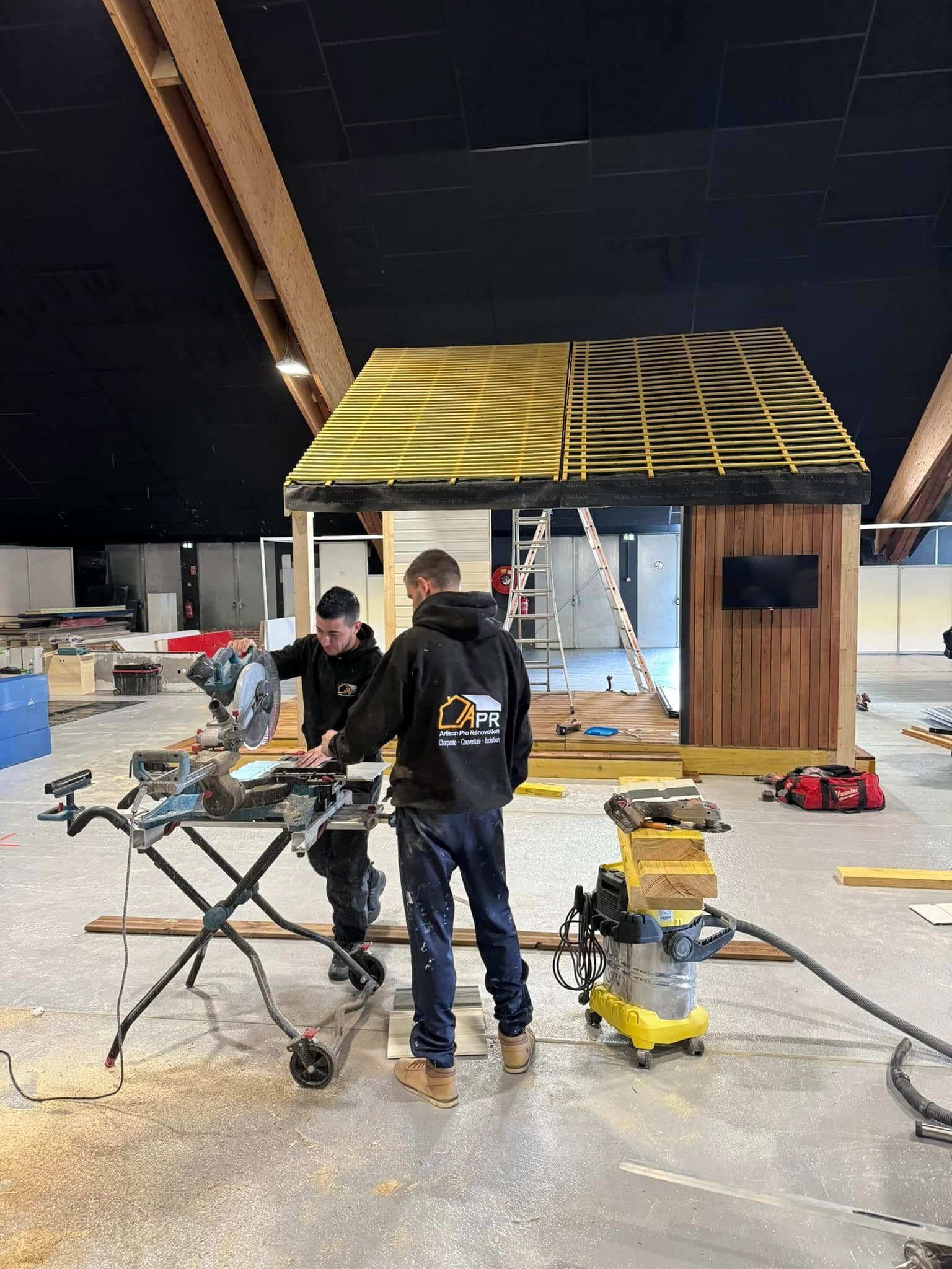 Deux ouvriers construisent une petite structure en bois, l'un à la scie. Présence de sciure au sol.