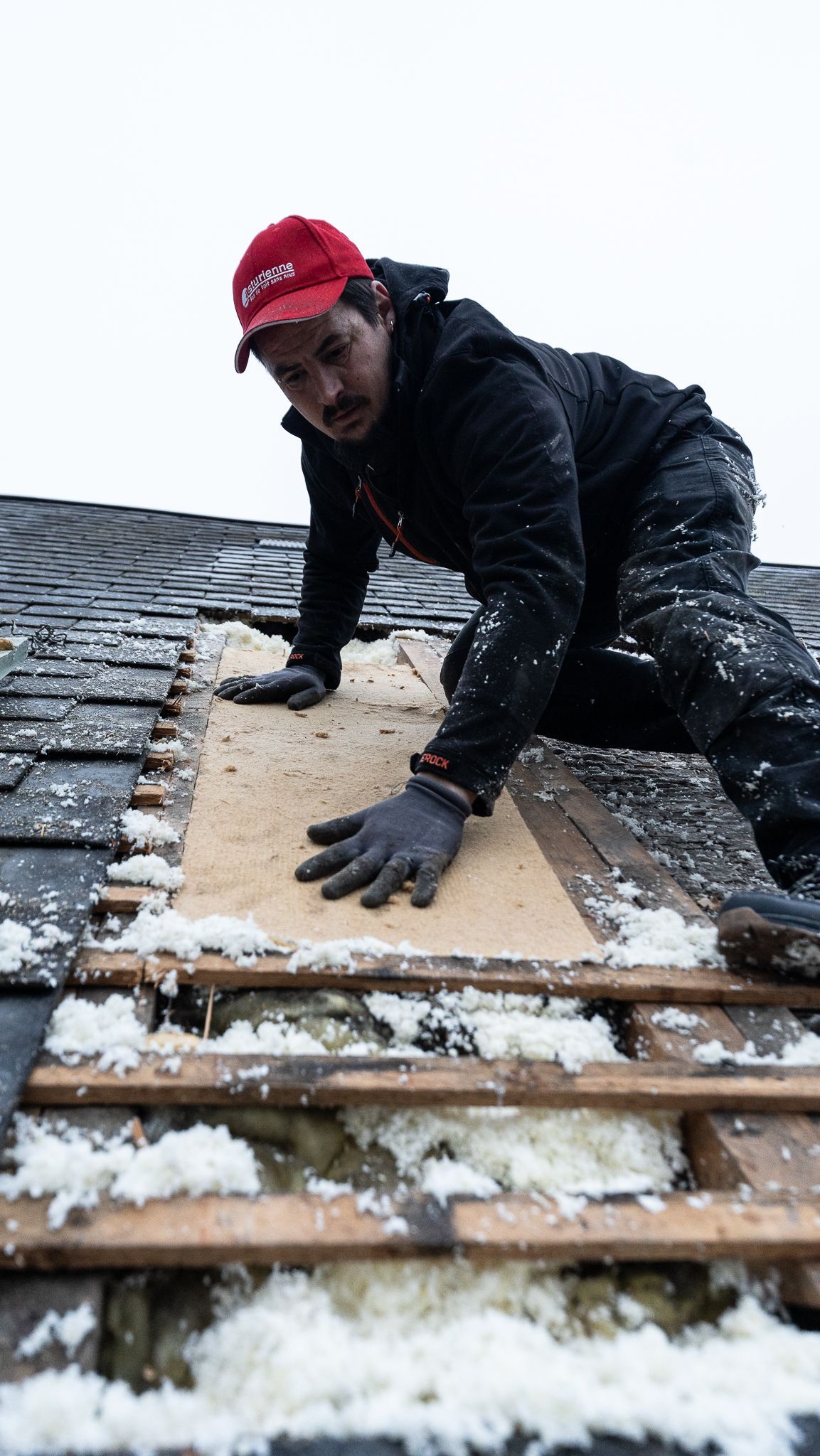 Couvreur en veste noire et casquette rouge remplaçant des planches de toiture, travaillant par temps nuageux.