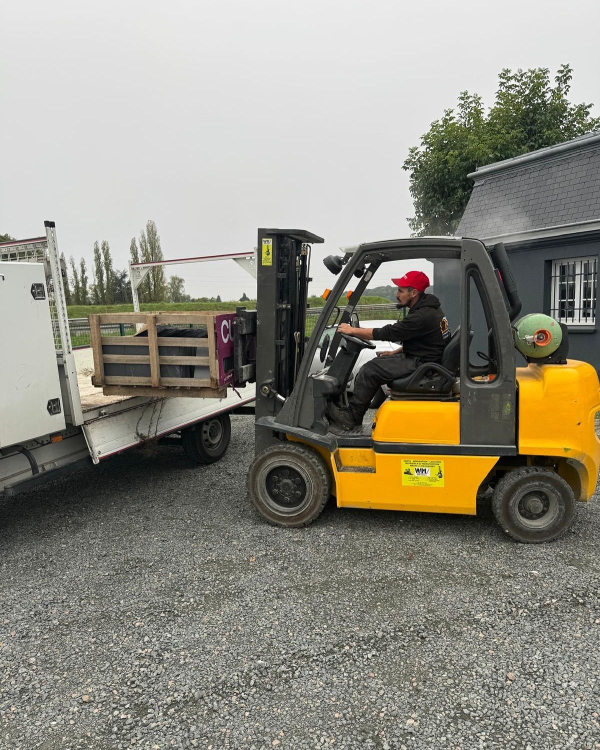 Un homme portant une casquette rouge conduit un chariot élévateur jaune et charge une caisse sur un camion à l'extérieur.