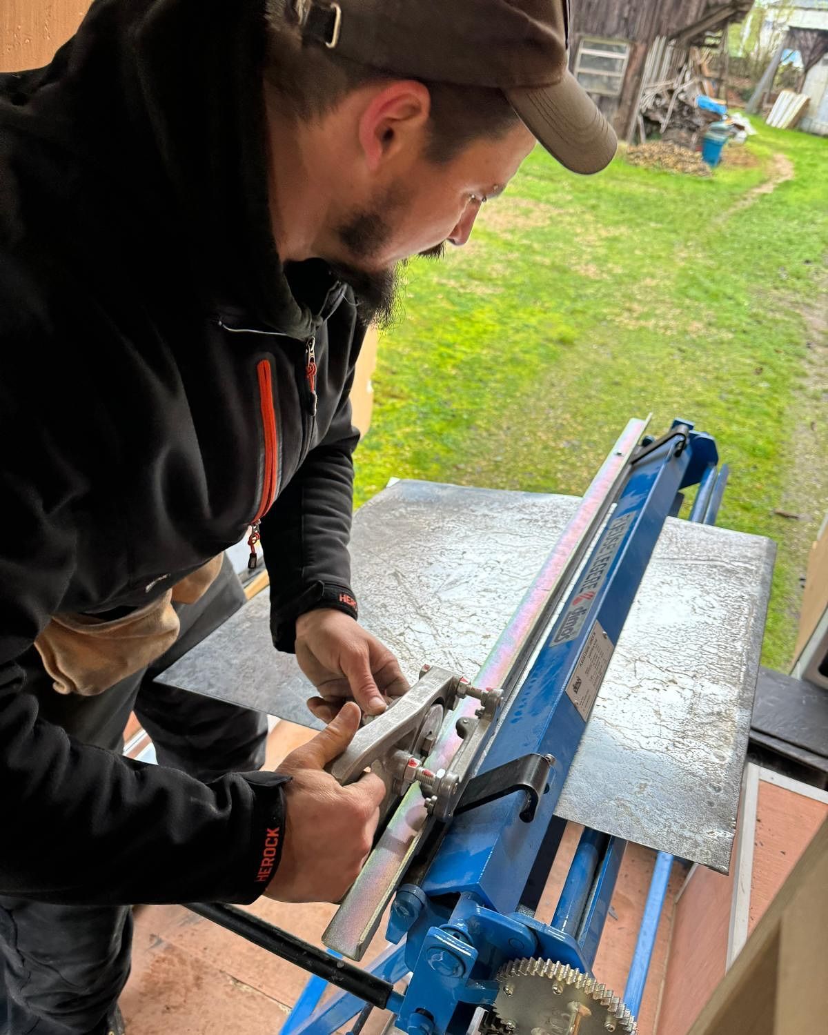Un homme utilise une cisaille à métal bleu pour couper une feuille de métal argenté à l'extérieur, portant une casquette et une veste noire.