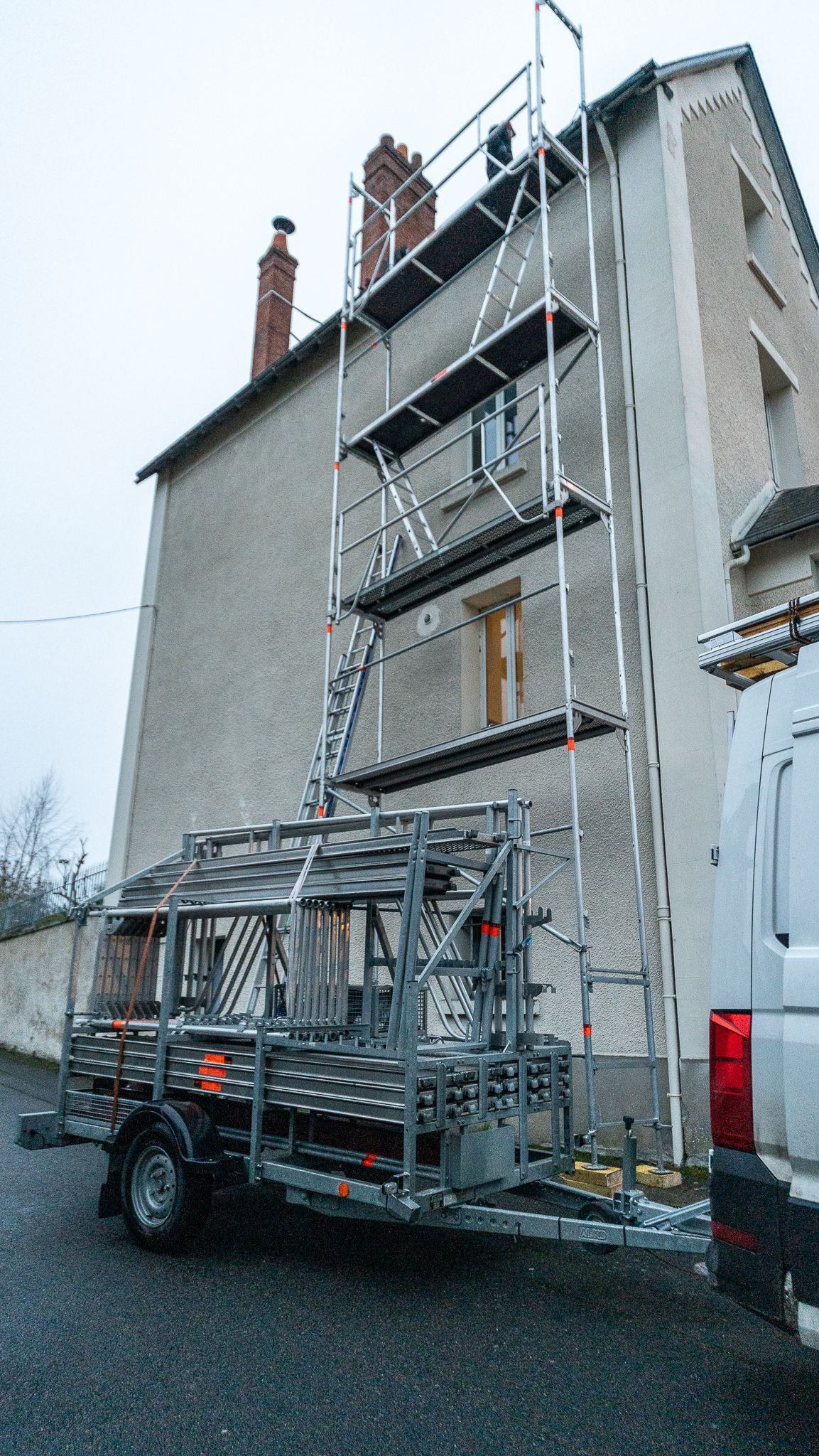Échafaudage assemblé contre un bâtiment, avec une remorque chargée de composants au premier plan. Extérieur gris.